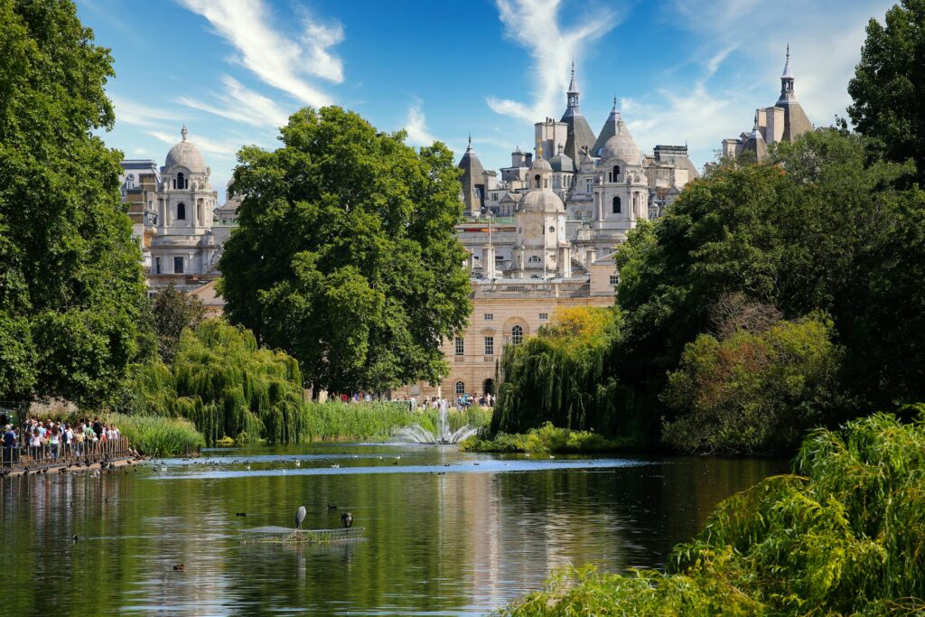 A scenic view of Hyde Park in London with a lake, birds resting on the water, many trees, a fountain, and a crowd walking nearby